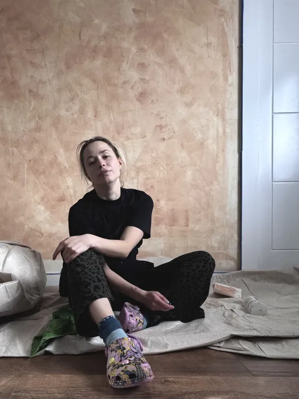 Vera Hylands painting a mural in Belfast — sitting in front of a warm terracotta wall, work in progress